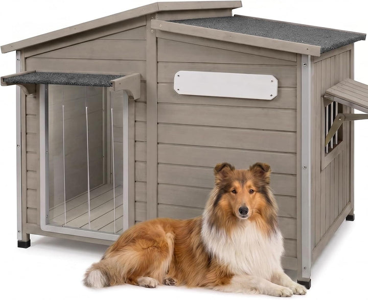 A brown and white collie dog lies in front of a large gray wooden dog house with a slanted roof, and small window, 