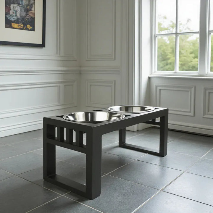  elevated pet feeder table with two stainless steel bowls, set on a gray tiled floor in a bright room with white paneled walls and a window.